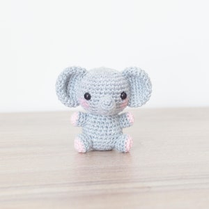 May include: A small grey crocheted elephant with pink accents on the ears and feet. The elephant has black eyes and a small grey trunk.