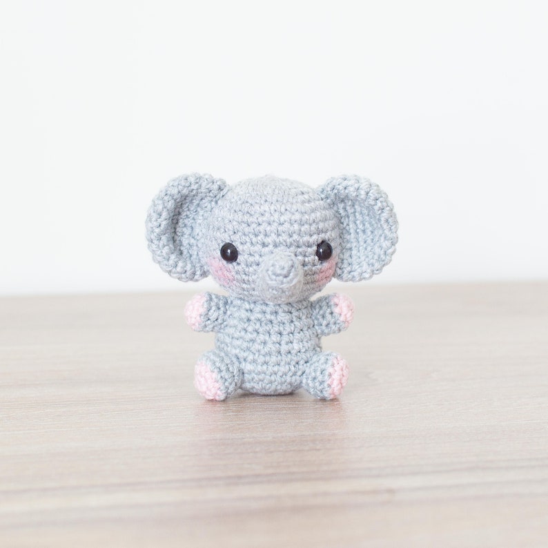 May include: A small grey crocheted elephant with pink accents on the ears and feet. The elephant has black eyes and a small grey trunk.