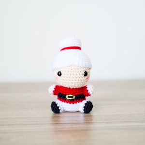 May include: A crocheted Mrs. Claus doll with a white hat and a red and black outfit. The doll has a cream-colored face with black eyes and rosy cheeks. The doll is sitting on a wooden surface.