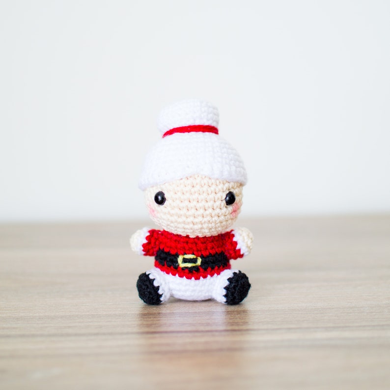 May include: A crocheted Mrs. Claus doll with a white hat and a red and black outfit. The doll has a cream-colored face with black eyes and rosy cheeks. The doll is sitting on a wooden surface.
