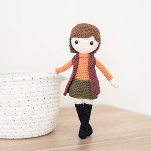 May include: A crocheted doll with brown hair, wearing a brown vest, orange sweater, green skirt, and black boots. The doll is standing on a wooden surface next to a white wicker basket.