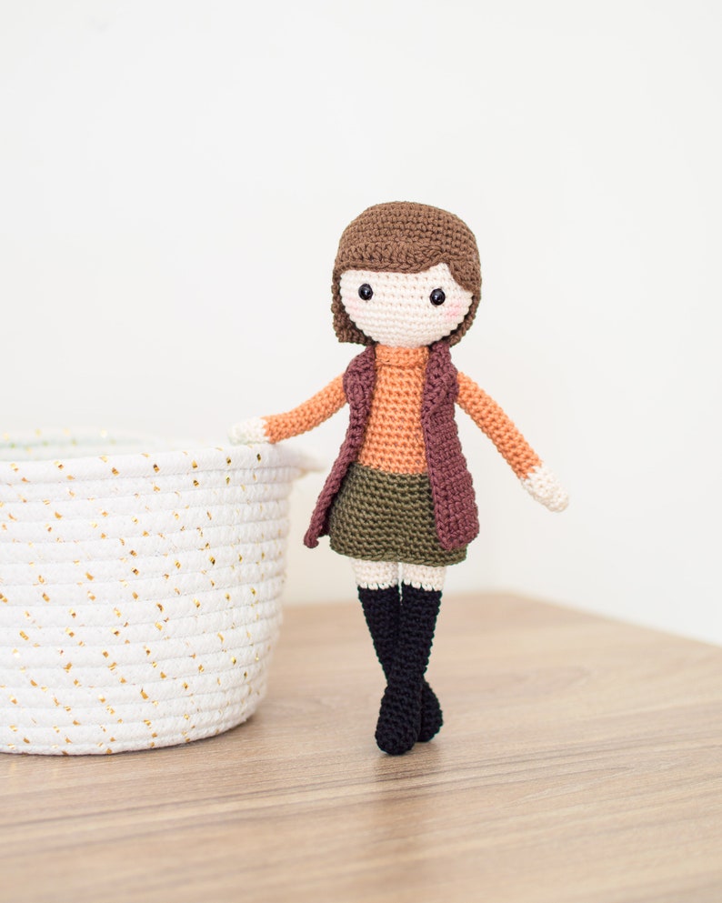May include: A crocheted doll with brown hair, wearing a brown vest, orange sweater, green skirt, and black boots. The doll is standing on a wooden surface next to a white wicker basket.