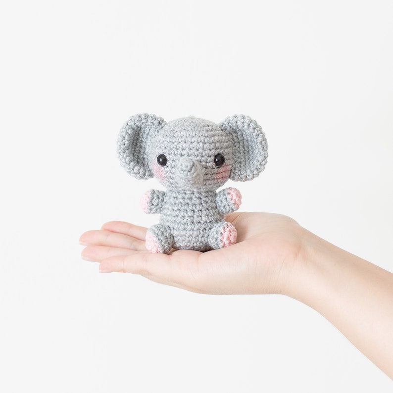 May include: A small grey crocheted elephant toy with pink accents on the ears and feet. The elephant is sitting in the palm of a hand.
