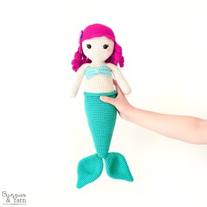 Op de afbeelding: Een gehaakte zeemeerminpop met roze haar, een blauw topje en een turquoise staart. De pop wordt vastgehouden door een hand.