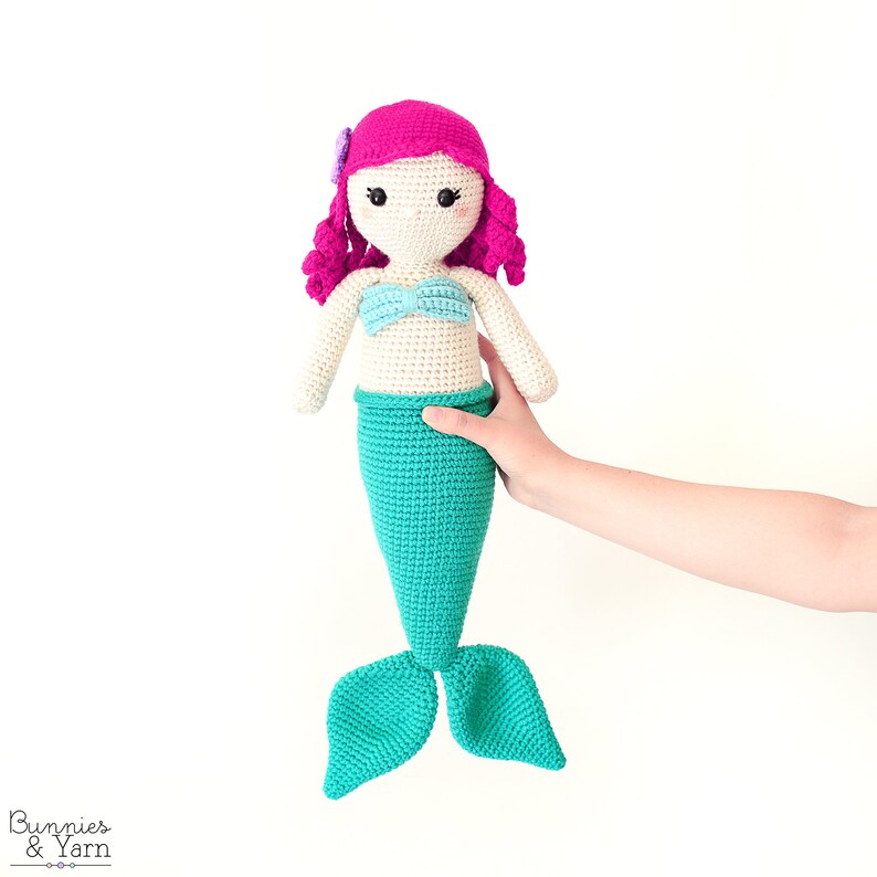 Op de afbeelding: Een gehaakte zeemeerminpop met roze haar, een blauw topje en een turquoise staart. De pop wordt vastgehouden door een hand.