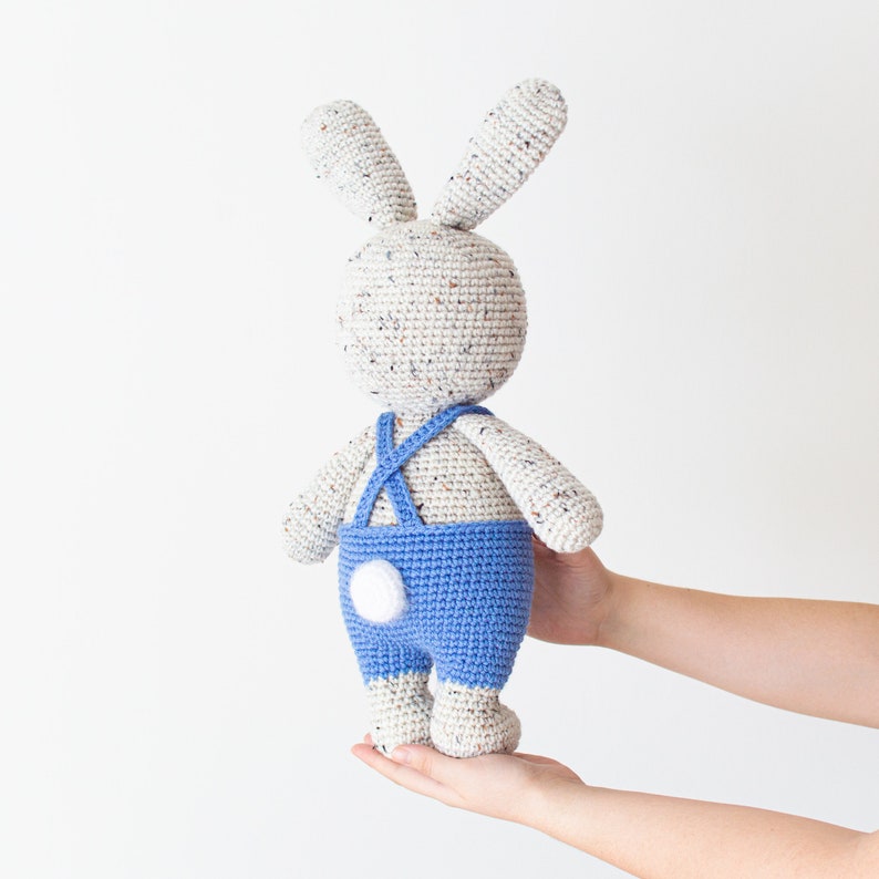 May include: A crocheted bunny toy wearing blue dungarees with a white button. The bunny is white with brown speckles and has long ears.