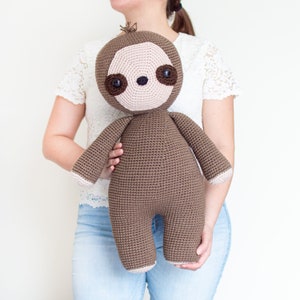 May include: A brown crocheted sloth plush toy with large brown eyes and a pink nose. The sloth is being held by a person wearing a white lace top and blue jeans.