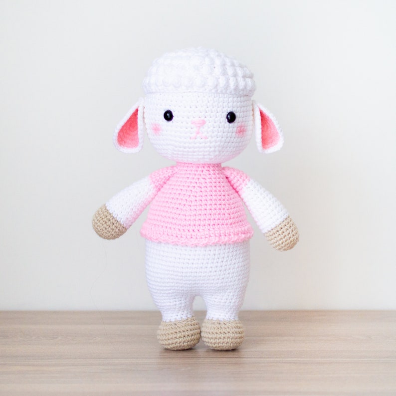 May include: A white crocheted lamb wearing a pink shirt. The lamb has a fluffy white head with black eyes and a pink nose. The lamb is standing on a wooden surface.