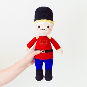 May include: A crocheted toy soldier with a red and yellow uniform, black boots, and a black hat. The toy has a white face with black eyes, a black mustache, and rosy cheeks.