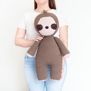 May include: A brown crocheted sloth plush toy with pink facial features and brown eyes. The sloth is being held by a person wearing a white shirt and blue jeans.