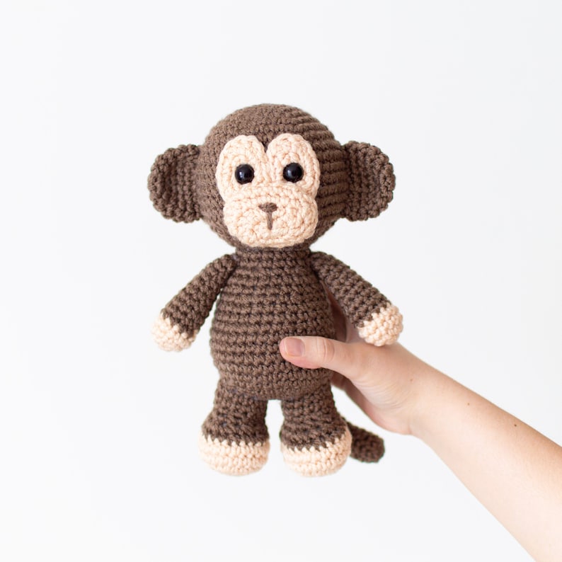 May include: A brown crocheted monkey toy with a pink face and black eyes. The monkey is standing on a white surface.