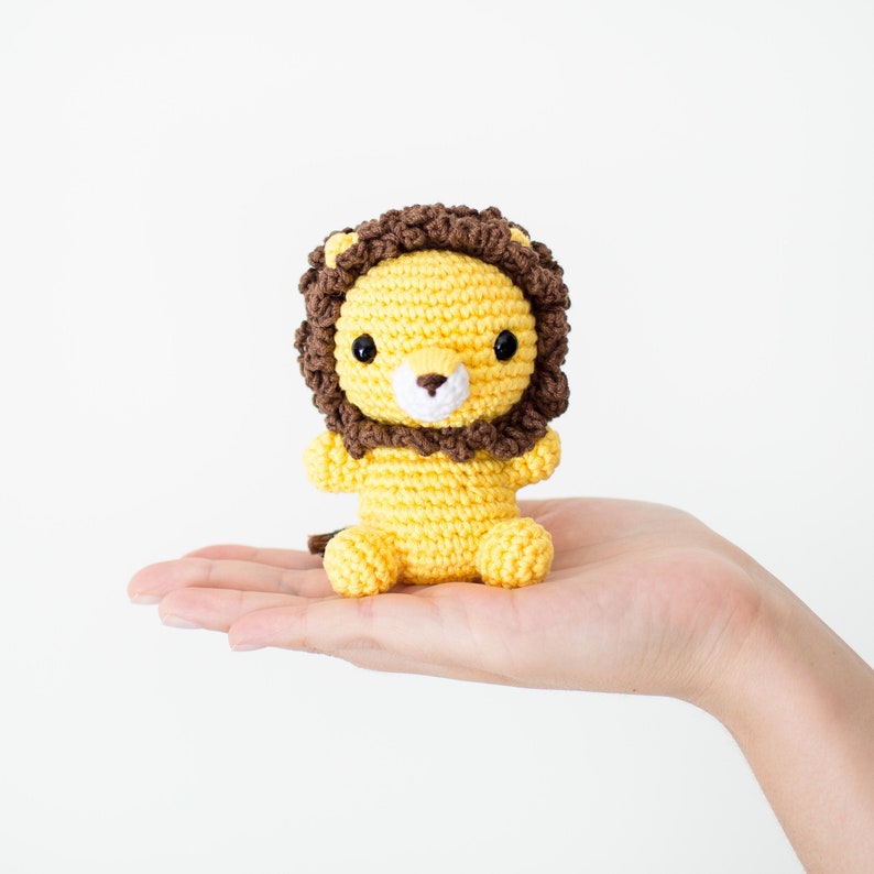 May include: A small crocheted lion toy with a brown mane, yellow body, and black eyes. The lion is sitting on a hand.