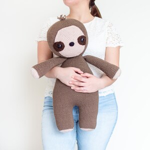 May include: A brown crocheted sloth toy with large brown eyes and a pink nose. The sloth is being held by a person wearing a white lace top and blue jeans.