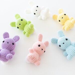 May include: Six crocheted bunny plushies in different colors: light blue, light pink, light green, purple, yellow, and white. The bunnies have black eyes and are lying on a white surface.