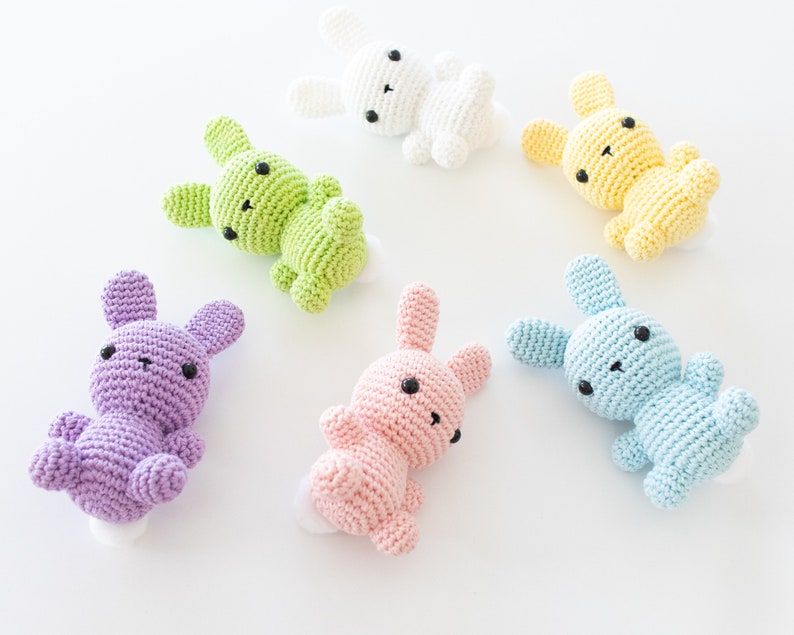 May include: Six crocheted bunny plushies in different colors: light blue, light pink, light green, purple, yellow, and white. The bunnies have black eyes and are lying on a white surface.