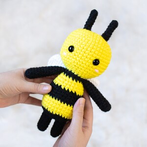May include: A crocheted bee plush toy with black and yellow stripes, white wings, and black eyes. The bee is being held by a person's hands.