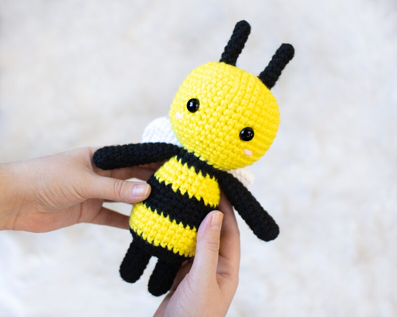 May include: A crocheted bee plush toy with black and yellow stripes, white wings, and black eyes. The bee is being held by a person's hands.