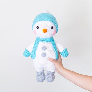 May include: A crocheted snowman toy with a blue hat, scarf, and mittens. The snowman has a white body, black eyes, an orange nose, and pink cheeks.