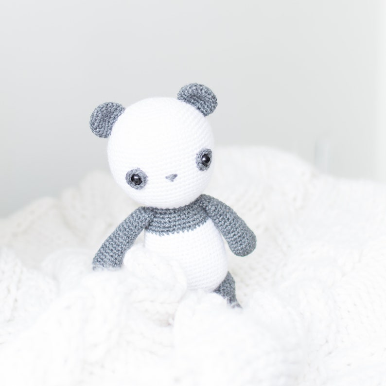 May include: A crocheted panda stuffed animal with gray ears and a gray band around its middle. The panda is sitting on a white fluffy blanket.