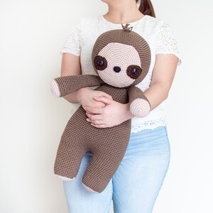 May include: A brown crocheted sloth toy with large brown eyes and a pink nose. The sloth is being held by a person wearing a white lace top and blue jeans.
