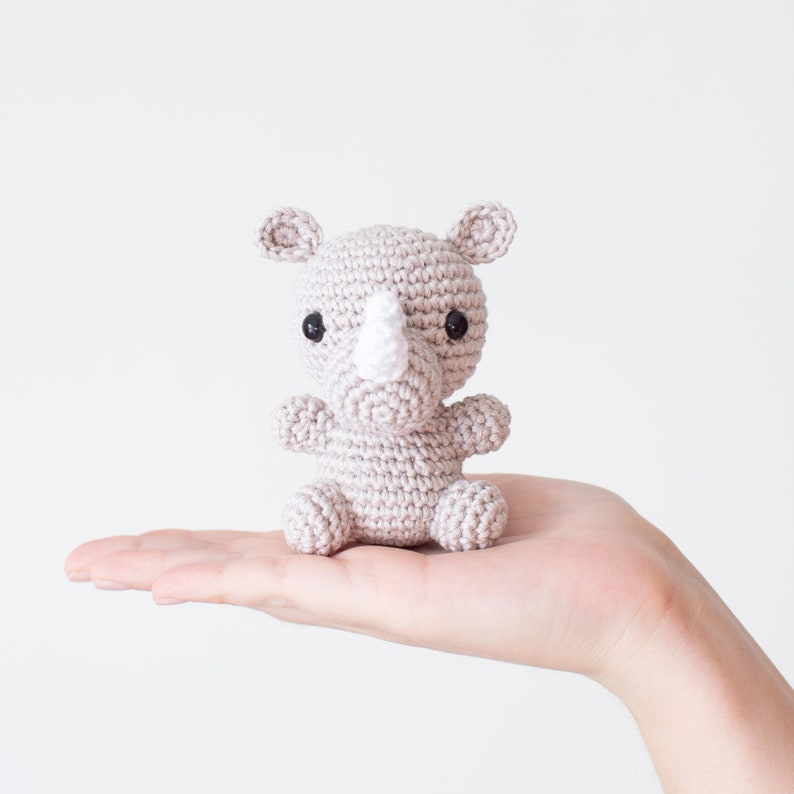 May include: A small crocheted rhinoceros toy in a light gray color with black eyes and a white horn. The toy is being held in a person's hand.