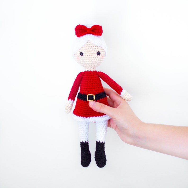 Puede incluir: Una mu&ntilde;eca de Navidad de crochet que lleva un vestido rojo con un cintur&oacute;n negro y ribete blanco, un sombrero blanco con un lazo rojo y botas negras. La mu&ntilde;eca est&aacute; siendo sostenida por una mano.