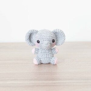 May include: A small grey crocheted elephant with pink accents on the ears and feet. The elephant has black eyes and a grey trunk.
