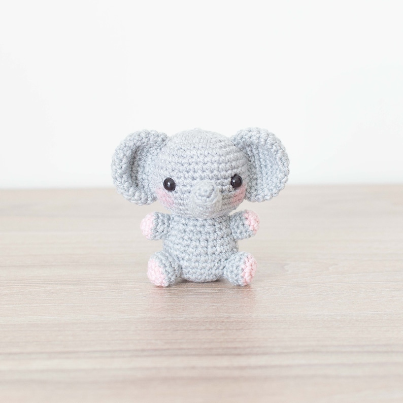 May include: A small grey crocheted elephant with pink accents on the ears and feet. The elephant has black eyes and a grey trunk.