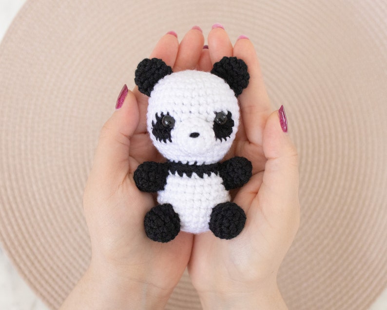 Op de afbeelding: Een klein gehaakt pandabeer speelgoed met zwarte oren, ogen en poten, en een wit lichaam. De panda wordt in de handpalmen van twee handen gehouden.