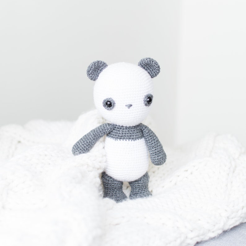 May include: A white crocheted panda bear with gray ears, arms, legs, and a band around its middle. The panda is sitting on a white crocheted blanket.
