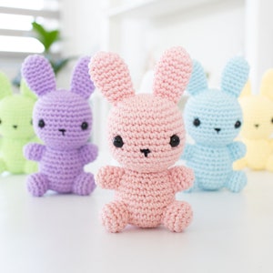 May include: Five crocheted bunny plushes in different pastel colors: green, purple, pink, blue, and yellow. The bunnies have black eyes and noses and are arranged in a row on a white surface.