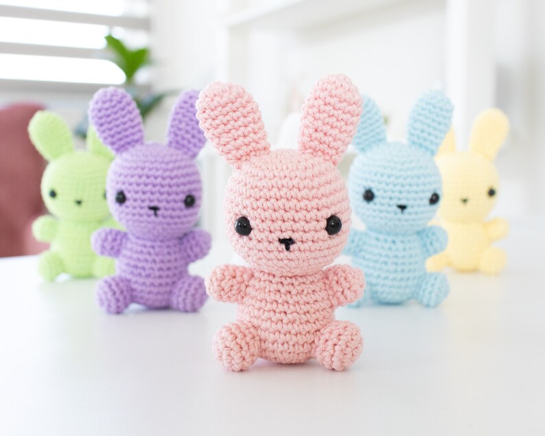 May include: Five crocheted bunny plushes in different pastel colors: green, purple, pink, blue, and yellow. The bunnies have black eyes and noses and are arranged in a row on a white surface.