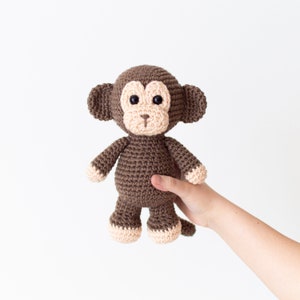 May include: A brown crocheted monkey toy with beige accents. The monkey has black eyes and a small pink nose.