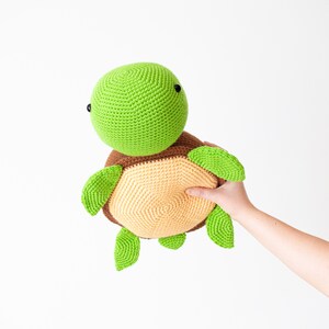 May include: A green and brown crocheted turtle toy with black eyes. The turtle is being held by a hand.