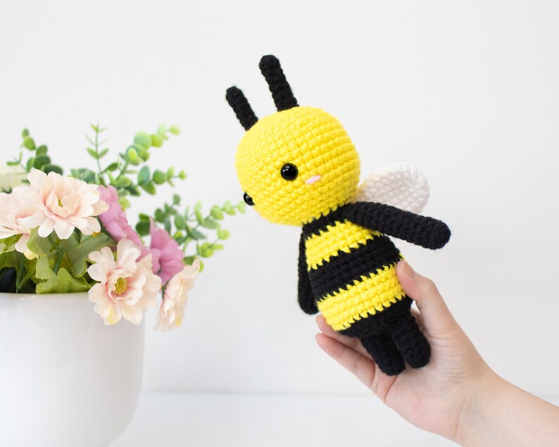 May include: A crocheted bee plush toy with black and yellow stripes, white wings, and black antennae. The bee has black eyes and a pink blush on its cheeks.