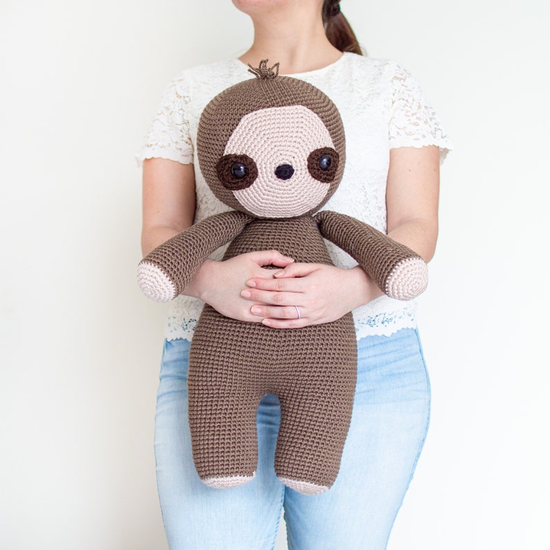 May include: A brown crocheted sloth plush toy with pink facial features and brown eyes. The sloth is being held by a person wearing a white lace top and blue jeans.