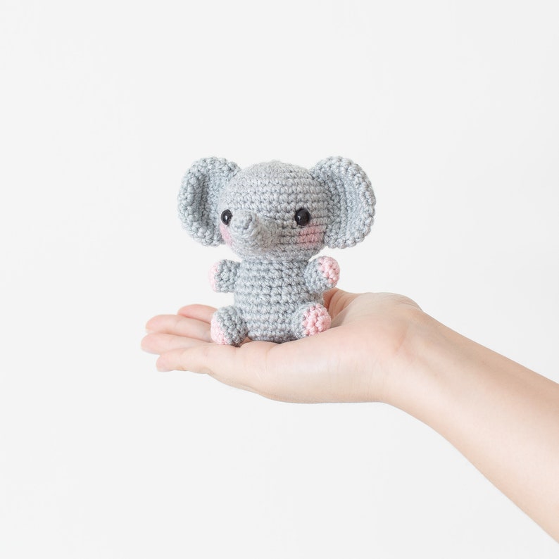 May include: A small grey crocheted elephant toy with pink accents on the ears and feet. The elephant is sitting in the palm of a hand.