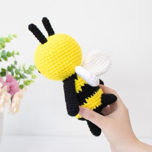 May include: A crocheted bee toy with black and yellow stripes, white wings, and black antennae. The bee is held in a hand.