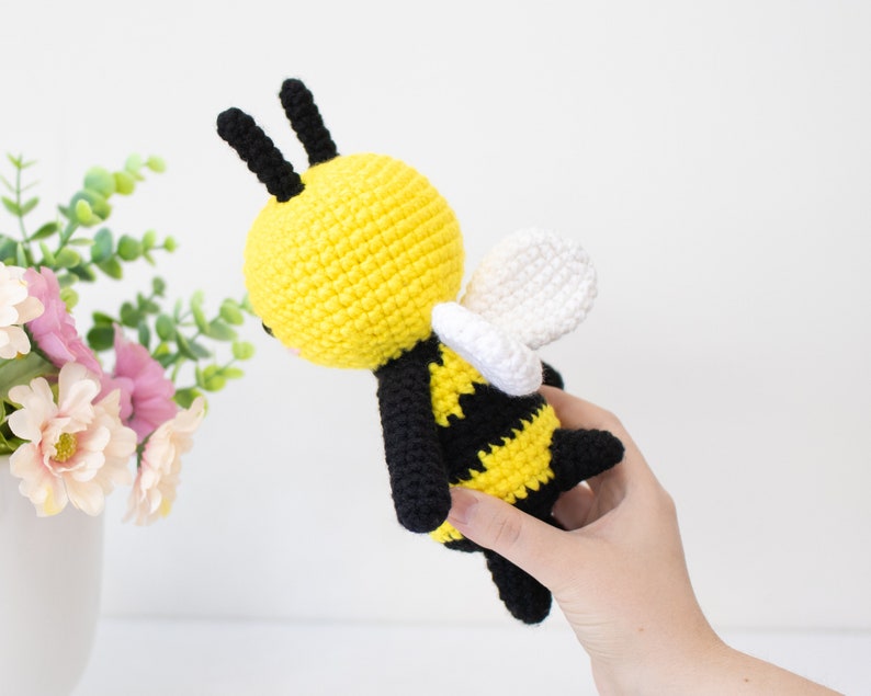 May include: A crocheted bee toy with black and yellow stripes, white wings, and black antennae. The bee is held in a hand.