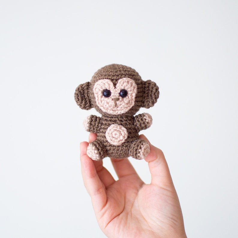May include: A small brown crocheted monkey toy with black eyes and a pink nose. The monkey is being held in a hand.