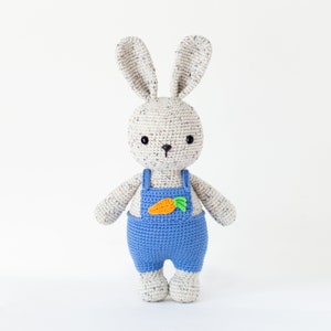 May include: A crocheted bunny toy wearing blue dungarees with a carrot patch. The bunny has white fur with brown speckles and black eyes.