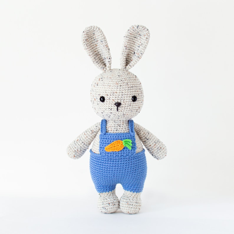 May include: A crocheted bunny toy wearing blue dungarees with a carrot patch. The bunny has white fur with brown speckles and black eyes.