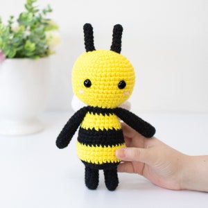 May include: A crocheted bee toy with black and yellow stripes, black legs, and black antennae. The bee has a yellow face with black eyes and a pink nose.