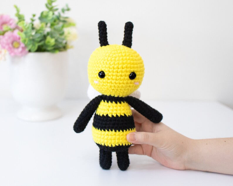 May include: A crocheted bee toy with black and yellow stripes, black legs, and black antennae. The bee has a yellow face with black eyes and a pink nose.