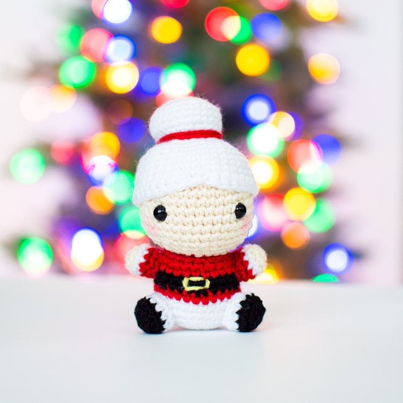 May include: A crocheted Santa Claus doll with a white hat and red trim. The doll has a beige face, black eyes, and a red and black outfit with a gold buckle. The background is a blurred display of colorful Christmas lights.