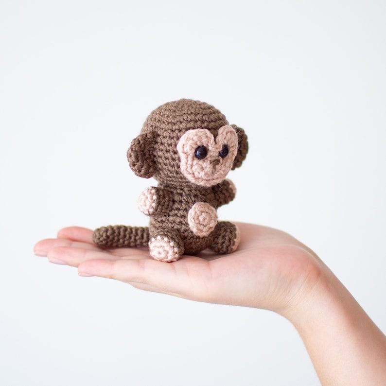 May include: A small brown crocheted monkey toy with pink facial features. The monkey is sitting on an outstretched hand.