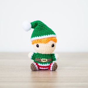 May include: A crocheted elf doll with a green hat, white trim, and orange hair. The elf is wearing a green and white striped shirt with a brown belt and red and white striped pants.