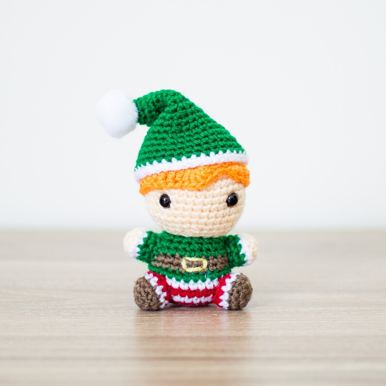 May include: A crocheted elf doll with a green hat, white trim, and orange hair. The elf is wearing a green and white striped shirt with a brown belt and red and white striped pants.