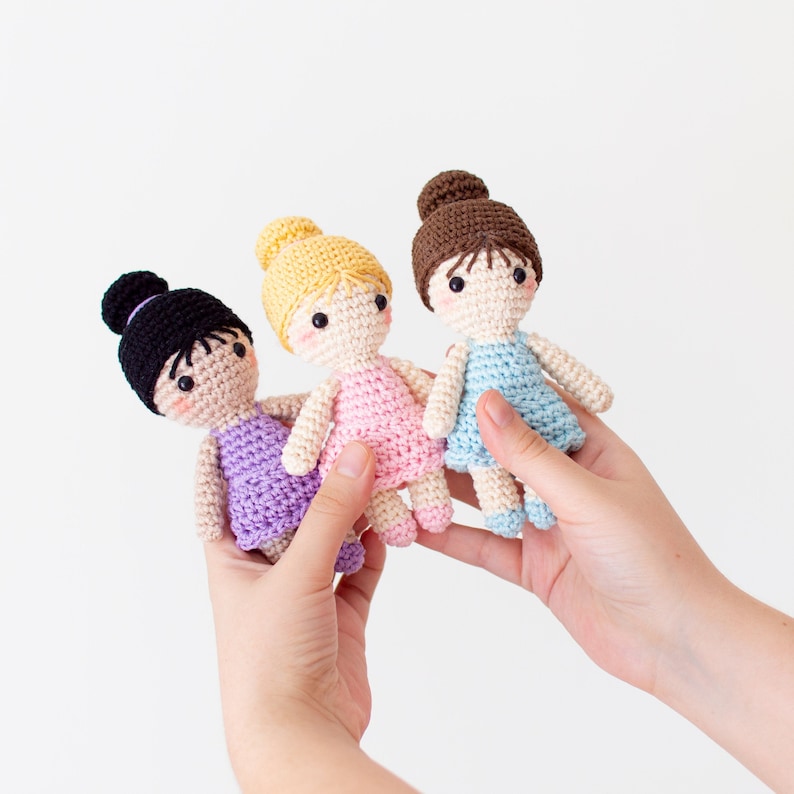 May include: Three crocheted dolls with different colored hair and dresses. The dolls are wearing pink, purple, and blue dresses. The dolls are being held by hands.