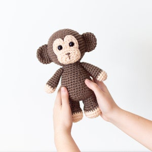 May include: A brown crocheted monkey toy with beige accents on the face and feet. The monkey is being held by two hands.